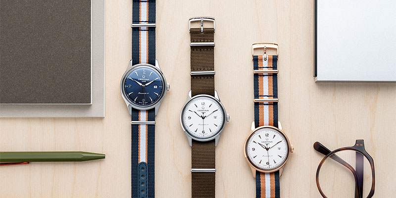 3 stycken Certina klockor med tygband liggandes på ett ljust bord. Två klockor med vita urtavlor och en med blå. 
