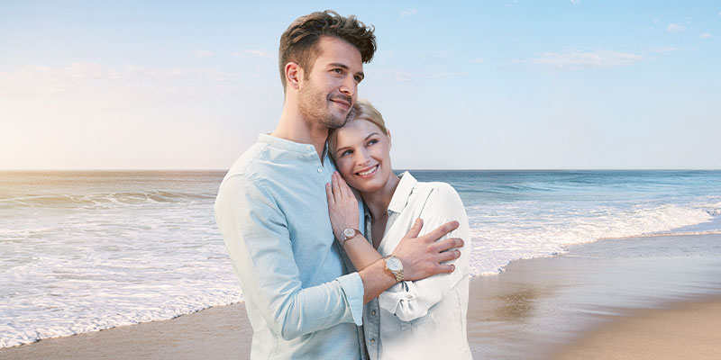 2 guldiga Certina klockor på en tjej och en kille som håller om varandra på en strand med havet och en blå himmel i bakgrunden.