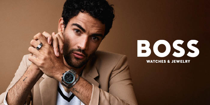Brunhårig man mot en en brun bakgrund som håller upp sina händer. På handleden har han en klocka från Boss och i bakgrunden står en vit text Boss, watches and jewelry.