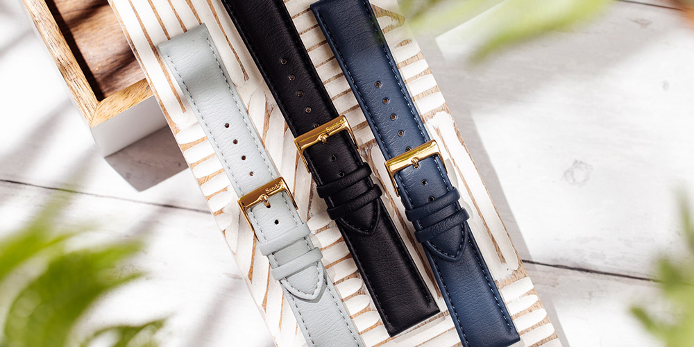 En grupp av tre klockarmband från Sandell, i färgerna ljusblå, svart och mörkblå, ligger diagonalt på en räfflad, ljus träyta. Armbanden har guldspännen. Delar av gröna blad syns suddigt i förgrunden och bakgrunden, vilket ger bilden en mjuk inramning.
