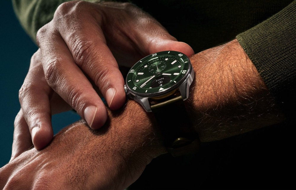 En man med grön tröja håller i boetten på sin gröna smartwatch från Withings. Klockan har ett traditionellt, analogt utseende trots sina smarta funktioner.
