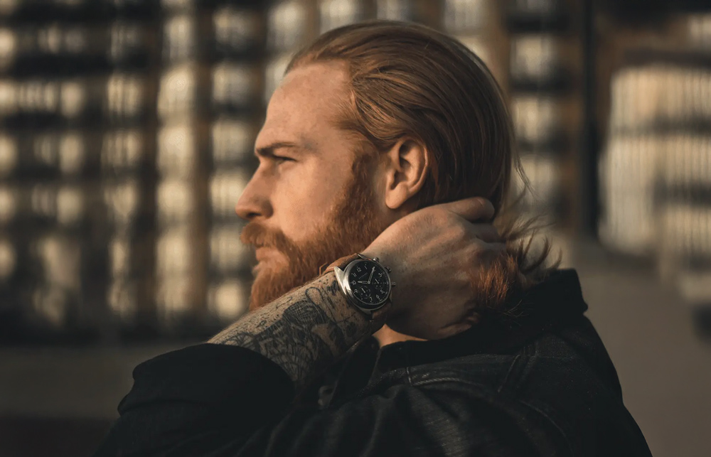 En man med långt hår och skägg håller sig i bakhuvudet och visar upp sin sportiga smartwatch från Garmin – en klocka med smarta funktioner för både vardagsliv och träning.