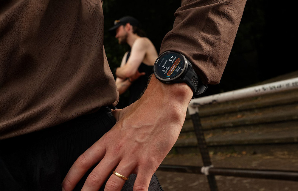 En man i brun tröja bär en svart sportklocka från Garmin på handleden. Han står i en mörk miljö framför ett stängsel med sina vänner – en scen som förmedlar styrka, gemenskap och funktionell stil.